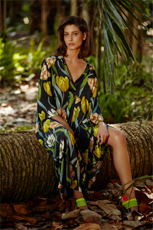 Model sitzt im Dschungel auf Baumstamm und trägt langes lässiges Kleid in schwarz mit gelben Tulpen von der Designermarke Alembika